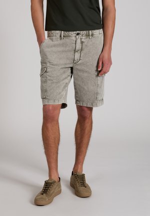 Mężczyzna w jasnoszarych spodenkach typu cargo, oliwkowo-zielonych trampkach i ciemnozielonej koszulce, stoi z jedną ręką w kieszeni.