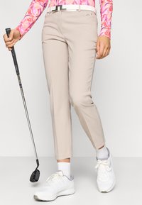 Μπεζ παντελόνια γκολφ με στενή εφαρμογή, με λείο υφή, λευκή ζώνη και έναν γκολφέρ που κρατάει μια ρακέτα, συνδυασμένα με λευκά αθλητικά παπούτσια.