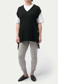 Svart stickad väst med V-hals över en vit T-shirt, kombinerad med grå avsmalnande byxor och svarta slip-on skor. Utsmyckad med sidoknyten.