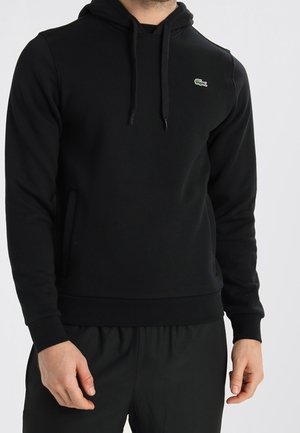 Man wearing a black hooded sweatshirt with a small green crocodile logo on the left chest and matching black pants.