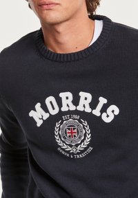 Homme portant un pull bleu marine foncé avec le texte blanc "MORRIS" et un emblème représentant un drapeau Union Jack et une couronne de laurier sur la poitrine.
