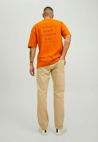 Oranžs kokvilnas T-krekls ar drukātu tekstu aizmugurē, apvienots ar gaiši beigu biksēm un baltām kedām ar tumšu svītru.