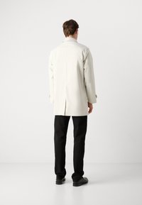 Abrigo blanco largo con abertura trasera y puños abotonados, llevado sobre pantalones y zapatos negros. Textura suave, diseño minimalista, colores neutros.