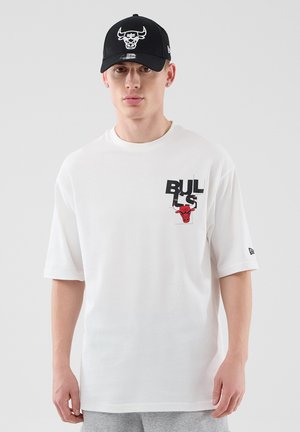 Young man wearing white Chicago Bulls T-shirt with black and red logo and black cap featuring white Bulls logo, standing against plain background.