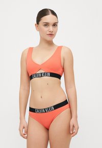 Koralo spalvos raukšlėtas bikini komplektas su trikampio formos liemenėle ir derančiomis aukštomis kelnaitėmis, papuoštas juodu "CALVIN KLEIN" logotipo juostele.
