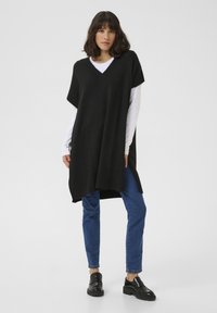 Zwarte gebreide poncho met een V-hals, korte mouwen en zijsplitten, gedragen over een witte lange mouwen shirt en gecombineerd met blauwe jeans.