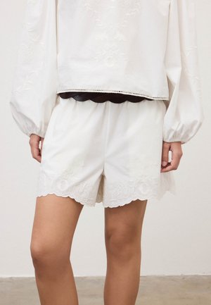 Person iført hvide broderede shorts med bølgekantede kanter og en hvid langærmet top med et sort fletbælte i taljen.