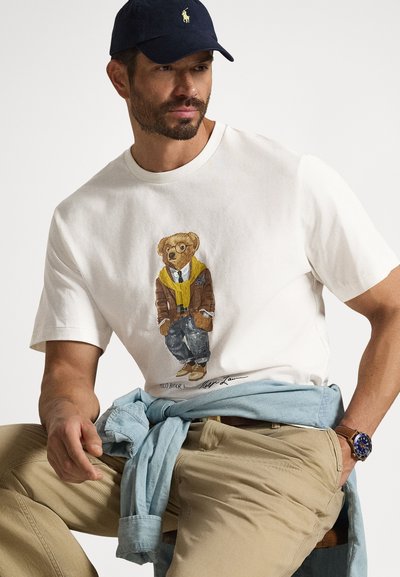 Άνδρας που φοράει λευκό μπλουζάκι με γραφικό ενός αρκούδου σε καφέ μπουφάν, μπεζ παντελόνι, μπλε καπέλο, ρολόι, και ανοιχτό μπλε πουκάμισο δεμένο γύρω από τη μέση.