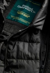 Sort polstret jakke med en synlig grøn etiket, der læser "Portland Outerwear Co. Outdoor Premium Quality Goods", syet indeni kraveområdet.