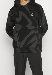 Personne portant un sweat à capuche à motifs abstraits noir et gris foncé avec un logo Jumpman blanc et un pantalon cargo noir, se tenant les mains dans le dos.