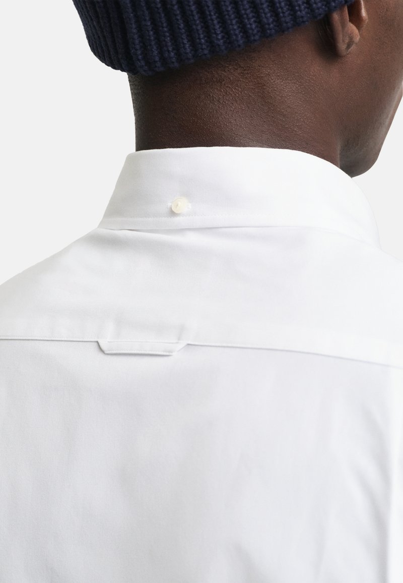 Vue arrière d'une personne portant une chemise blanche à boutons et un bonnet en tricot bleu marine, mettant l'accent sur le col de la chemise et le haut du dos.