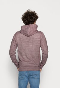 Persona con cabello rizado que lleva una sudadera marrón y jeans azules, de pie con las manos en los bolsillos, mirando hacia atrás contra un fondo liso.