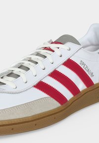 Λευκό αθλητικό παπούτσι Adidas με κόκκινες ρίγες, λεπτομέρεια από μπεζ σουέτ, λευκά κορδόνια, σόλα από καουτσούκ και σήμανση "SPEZIAL" στο πλάι.