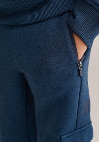 Mörkblå joggerbyxor i texturerad tyg, med sidodragkedjefickor och en präglad logotyp på benet.