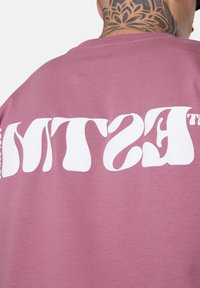 Person mit Nackentattoo, der ein mauvefarbenes Shirt trägt, auf dem große weiße verzerrte Schrift über den oberen Rücken aufgedruckt ist.