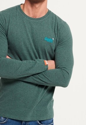 Grünes langärmliges T-Shirt aus weichem Stoff, mit rundem Ausschnitt und einem kleinen blauen Logo auf der Brust, mit einem Etikett am Ärmel.