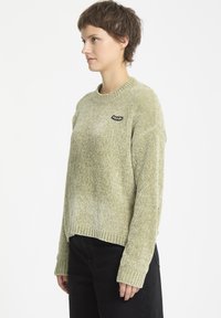 Pull vert clair avec un tricot texturé, poignets et ourlet côtelés, et un petit logo noir sur la poitrine gauche.