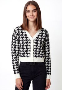 Zwart-witte vest met pied-de-poule patroon, V-hals, knopen aan de voorkant en geribbelde boorden, gecombineerd met een zwarte broek.