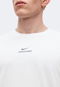 Άνδρας που φοράει λευκό μπλουζάκι Nike Pro με στρογγυλή λαιμόκοψη, που δείχνει το πηγούνι, το λαιμό και το κάτω μέρος του προσώπου σε απλό φόντο.