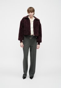 Junge Person, die eine lila kuschelige Jacke, ein cremfarbenes Strickoberteil, graue Hose mit einem Gürtel mit goldener Schnalle und schwarze spitze Schuhe trägt und vor einem weißen Hintergrund steht.