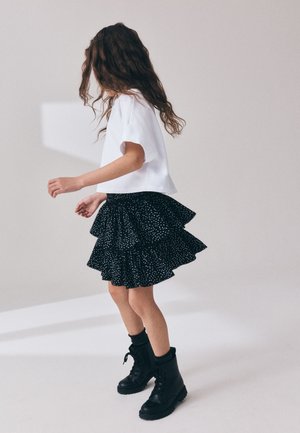 Junges Mädchen mit lockigem Haar, trägt ein weißes kurzärmliges Oberteil, schwarzen mehrlagigen Rock mit Punkten, schwarze Socken und schwarze Schnürstiefel, steht seitlich.