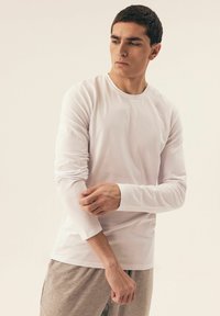 Langärmeliges weißes T-Shirt aus glattem Stoff, mit Rundhalsausschnitt und schmalen Bündchen, kombiniert mit hellgrauen Hosen.