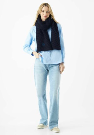 Écharpe en tricot bleu marine avec une texture côtelée, portée sur une chemise bleu clair. Présente un petit logo dans le coin. Coupe décontractée avec un jean à jambe droite.