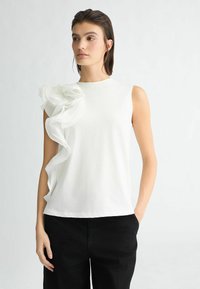 Top bianco senza maniche con un design di tessuto arricciato sulla spalla sinistra, realizzato in una texture liscia. Abbinato a pantaloni neri a gamba larga.