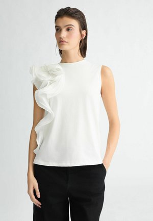 Top bianco senza maniche con un design di tessuto arricciato sulla spalla sinistra, realizzato in una texture liscia. Abbinato a pantaloni neri a gamba larga.