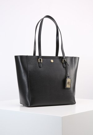 Black leather tote bag with dual shoulder straps, gold-tone hardware, and a hanging branded tag on a white surface.