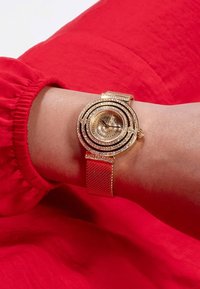 Zlatni mrežasti sat sa spiralnim dizajnom i kristalnim detaljima. Ima okruglu brojčanicu i teksturirani remen, leži na zapešću na crvenoj pozadini.