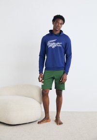 Marineblå hettegenser med krokodillegrafikk, kombinert med grønne shorts. Hettegenseren har kengurulomme og ribbede ermer. Myk, strukturert stoff.