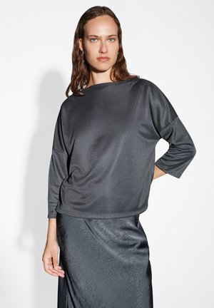 Frau mit nassen Haaren, die ein lockeres, dunkelgraues Oberteil mit drei Viertelärmeln und einen strukturierten, dunkelgrauen Rock vor einem weißen Hintergrund trägt.