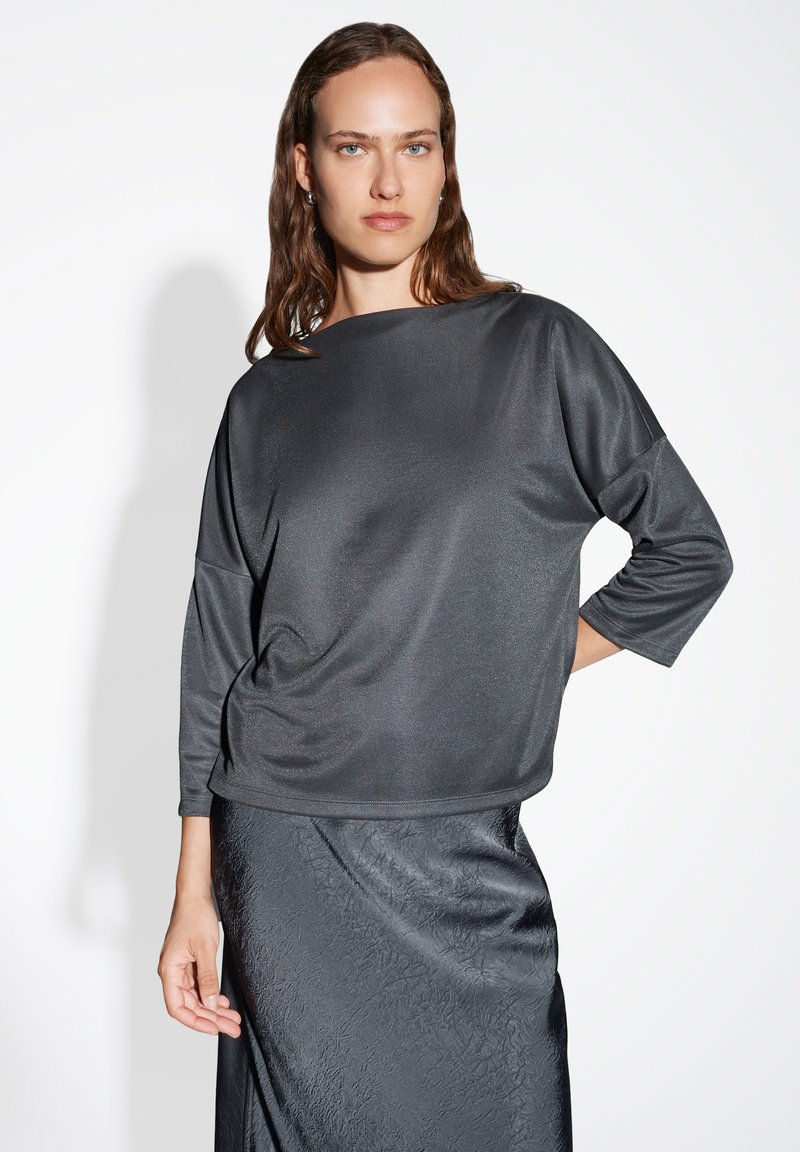 Femme aux cheveux mouillés portant un haut ample à manches trois quarts gris foncé et une jupe texturée grise foncée sur fond blanc.