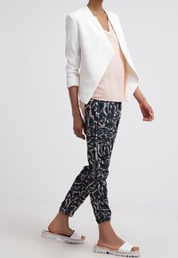 Blazer bianco su una maglietta rosa chiaro, abbinato a pantaloni jogger neri con motivo e sandali bianchi a scivolo. Caratteristiche includono una vestibilità comoda e un tessuto leggero.