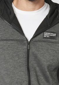 Homme portant un sweat à capuche gris à zip avec des bordures noires sur un t-shirt blanc col rond, affichant le texte « Koroshi Brand Urban Concept Est. 20.01 ».