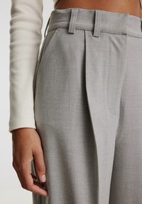 Lichtgrijze op maat gemaakte broek met plooien, gemaakt van een gestructureerde stof. Voorzien van een tailleband met riemlussen en zijzakken.