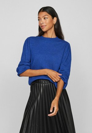 Femme aux longs cheveux foncés portant un pull bleu et une jupe plissée noire, posant les bras croisés et regardant sur le côté sur un fond gris.