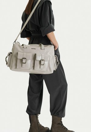 Osoba v černém overal a hnědých šněrovacích botách nese velkou béžovou kabelku přes rameno s několika předními a bočními kapsami.