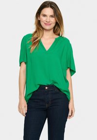 Grön kortärmad V-ringad blus i ett lätt material, med en avslappnad passform. Bärs ihop med mörka jeans, vilket visar en avslappnad stil.