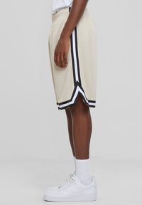 Mandige beige træningsshorts med sorte og hvide sidelister, lavet af mesh-stof, med elastisk talje og en løs pasform.