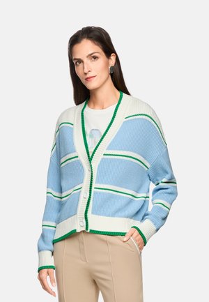 Woman wearing a blue and white striped cardigan with green trim over a white top and beige pants, standing with one hand in pocket.