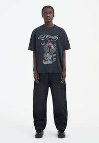 Černé grafické tričko s barevným designem ve stylu tetování, zobrazující lebku a křídla. Nosené s volnými černými džíny a černými botami.