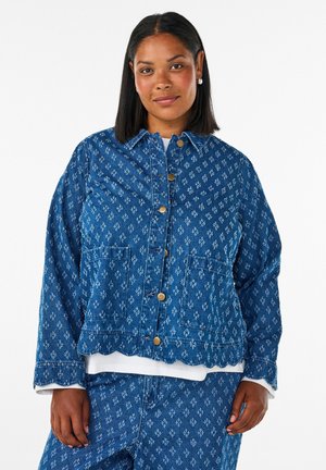 Frau mit glatten schwarzen Haaren trägt eine passende blaue gemusterte Jeansjacke und Hose mit goldenen Knöpfen und steht vor weißem Hintergrund.