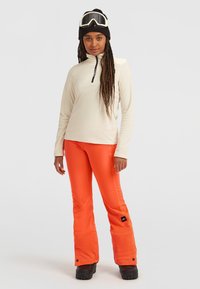 Crème kleurige fleece top met lange mouwen en een half-rits kraag, gecombineerd met feloranje geïsoleerde broek en zwarte snowboots. Sneeuwgoggles en een beanie inbegrepen.