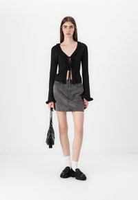 Top negru cu mânecă lungă, cu volănașe la margine și închidere cu fundiță, purtat cu o fustă din denim gri și pantofi negri, ținând o geantă mică neagră.