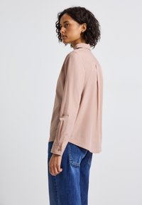 Femme avec des cheveux bouclés et foncés portant une chemise à manches longues rose clair et un jean bleu, tournée vers la gauche devant un fond blanc uni.