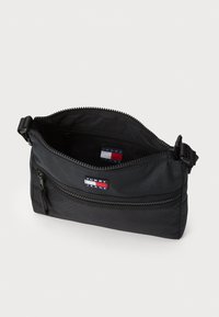 Sac bandoulière en tissu noir avec une fermeture éclair supérieure, deux compartiments, poche zippée extérieure et étiquette de logo. Texture lisse avec une forme rectangulaire.