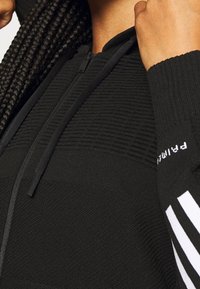 Sudadera con cremallera negra de tejido texturizado, con puños acanalados y capucha. Presenta acentos de rayas blancas en las mangas y un logotipo en el puño.