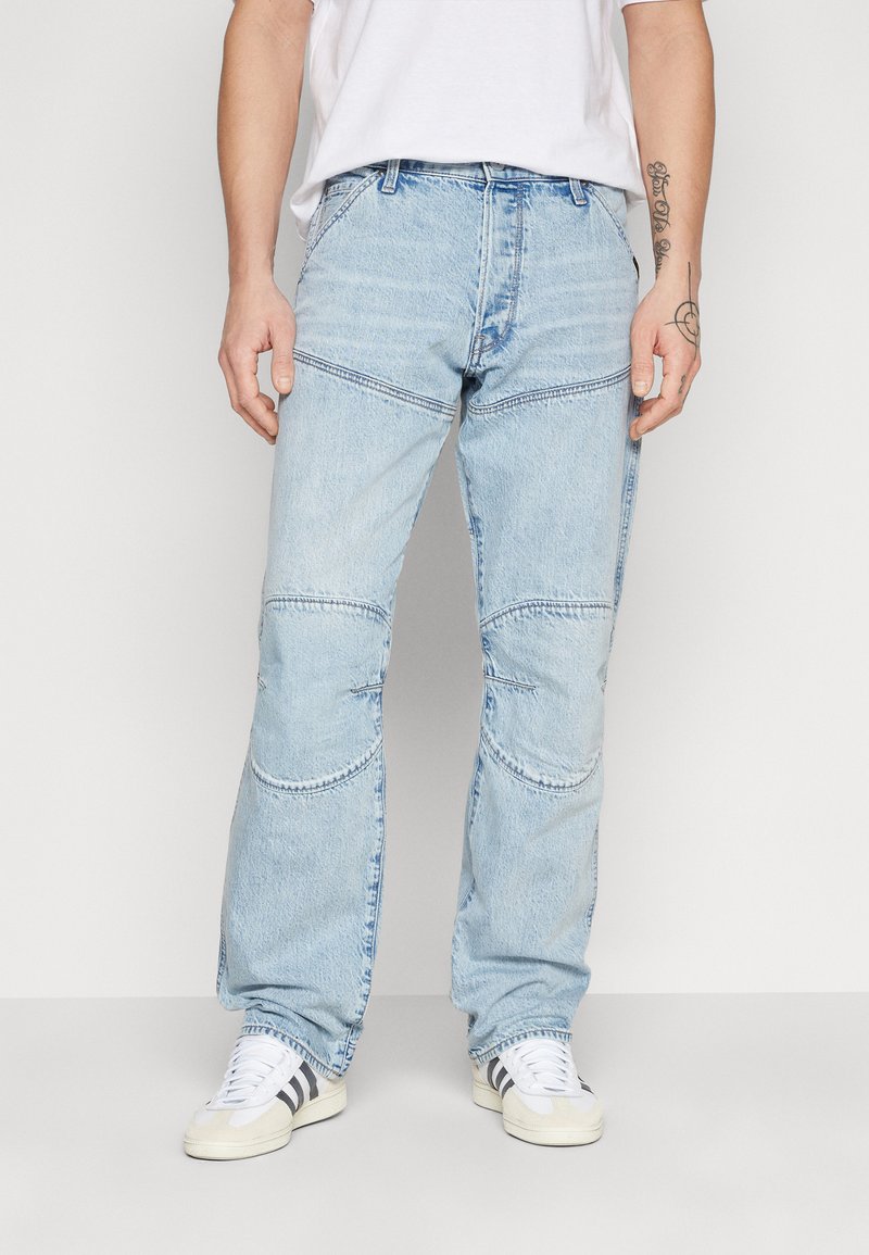 Lichtblauwe denim jeans met een losse pasvorm, patchwork-details en contrasterende stiksels, gecombineerd met witte sneakers.